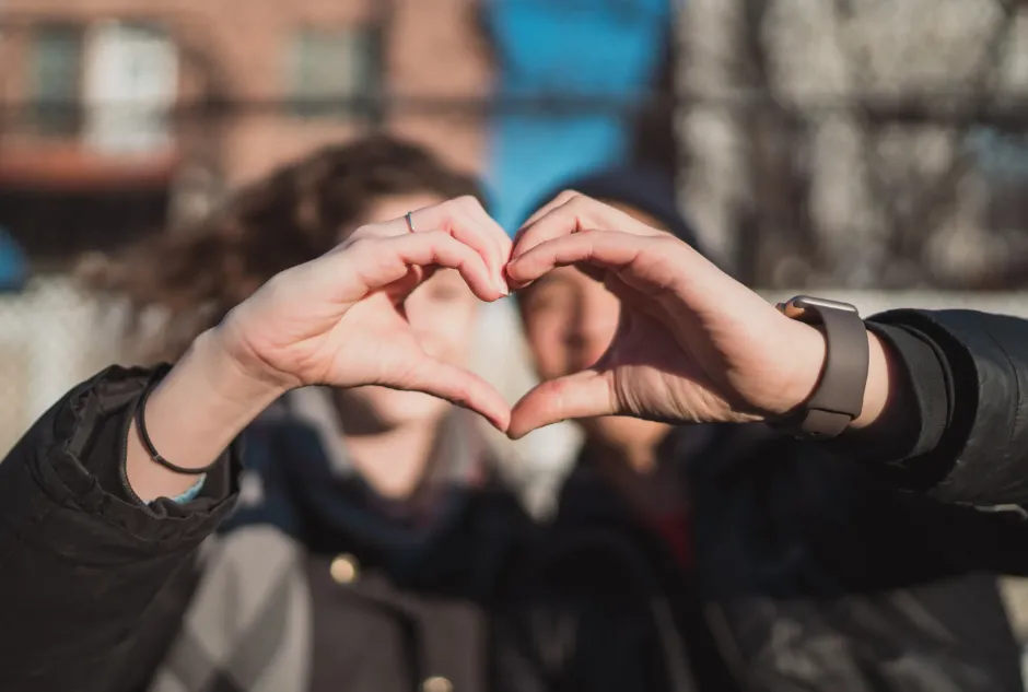  Two people stand side by side, their fingers forming a heart shape in the air