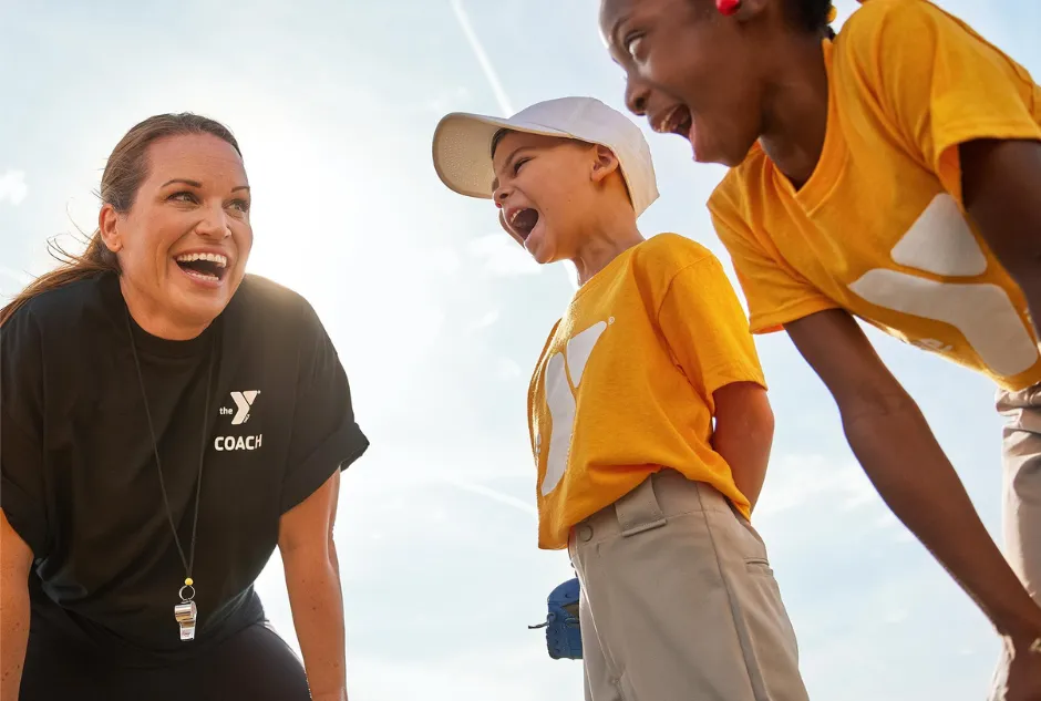 A female coach leans forward, hands resting on her knees, her face lit up with a warm smile as she encourages two young baseball players.