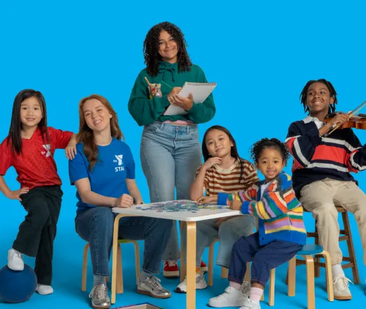 Two YMCA afterschool counselors and several children gathered around a table during an activity. One child is playing a violin while games and activity materials are spread across the table and floor.